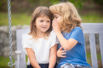 Wall Mural - Little boy whispers to lovely girl in ear. Brother and sister playing in spring park outdoors. Little boy and girl kids enjoying spring. Kids walking at backyard. Best friends two children.