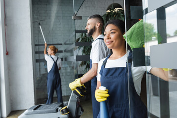 Wall Mural - happy bi-racial woman with rag and spray bottle near colleagues working in office.