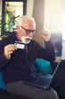 © Solid photos - Smiling senior man online shopping using credit card and laptop