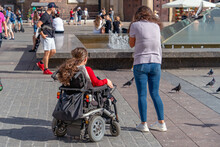 A Disabled Person In A Wheelchair On A Sunny Day On A City Street Among Healthy People Looks At The Fountain. Integration Of People With Disabilities Into Society