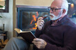 © Solid photos - Senior man smoking pipe and reading book by fireplace in winter