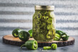 Jar of homegrown quick pickled homegrown jalapeno peppers surrounded by fresh green jalapeno peppers on a wooden cutting board