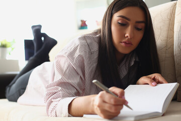 Portrait of smiling beautiful woman writing notes in notebook lying on sofa at home