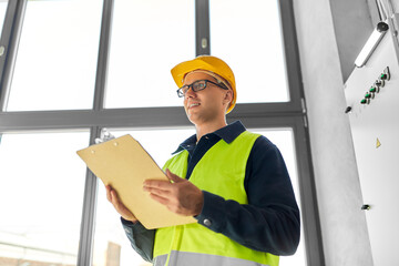 Sticker - construction business and building concept - happy smiling male electrician or worker in helmet and safety west with papers on clipboard and pencil at electric board