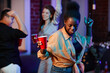 © Seventyfour - Group of young cheerful multicultural women in casualwear dancing at home party with dynamic black girl holding red cup in front