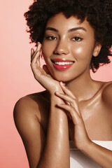 Vertical shot of African American girl with glowing, hydrated facial skin, white teeth, happy smile, apply facial moisturizer, pink background