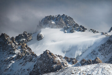 Wall Mural - A large rocky mountain with a rocky peak, on the mountain the Tuyuk-Su glacier is covered with deep snow, the sun is shining on the right, the sky is with dark thick clouds