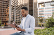 © Look! - Focused young african guy is using tablet sitting on street. Man with dark hair wears shirt. Technology concept, lifestyle