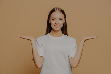 Wall Mural - Beautiful dark haired European woman raises palms pretends holding something over blank copy space against brown background wears casual white t shirt proposes product. Advertisement gesture