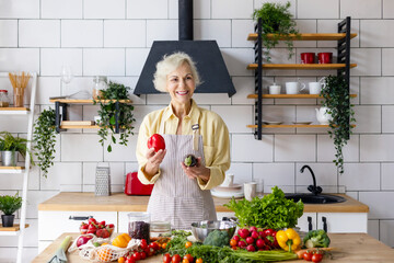 Wall Mural - beautiful elderly gray haired senior woman cook in cozy kitchen with fresh organic vegetables, tomatoes, cabbage, lettuce, cucumbers on table cooking healthy vegetable salad, healthy food active life