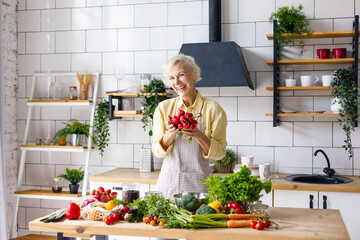 beautiful elderly gray haired senior woman cook in cozy kitchen with fresh organic vegetables, tomatoes, cabbage, lettuce, cucumbers on table cooking healthy vegetable salad, healthy food active life