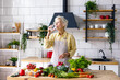 beautiful elderly gray haired senior woman cook in cozy kitchen with fresh organic vegetables on table for healthy vegetable salad, drink pure clear water from glass , healthy food for active life