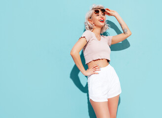 Wall Mural - Young beautiful smiling female in trendy summer shorts and top clothes. Sexy carefree woman posing near blue wall in studio. Positive blond model having fun and going crazy. Cheerful and happy