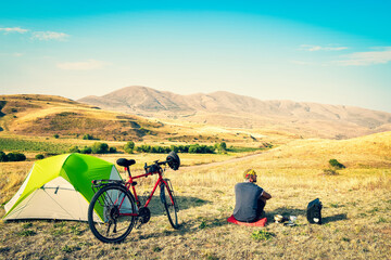 Caucasian man sit by touring bicycle in mountains on side of green tent have break time rest in morning. Solo travel journey. Long-term travel around the world