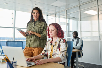 Poster - Young confident manager with tablet standing by female coworker with laptop while giving her advice about presentation of new project