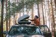 A young man opens the upper tourist trunk of a car in a pine forest on the shore of a lake at sunset.
