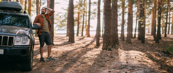 A young man stands next to a car with an upper tourist trunk in a pine forest on the shore of a lake at sunset.