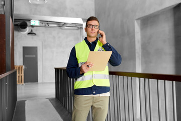 Wall Mural - architecture, construction business and building concept - male supervisor in safety west with clipboard calling on smartphone at office