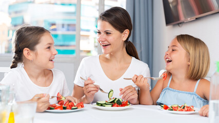 Wall Mural - Young mother and two daughters eating healthy food with vegetables in living room