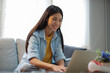 © ijeab - Young asian woman smiling and using laptop while sitting on couch at home.