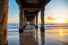 A Gorgeous Summer Landscape From Underneath The Manhattan Beach Pier With A Breathtaking Sunset In The Sky And Vast Blue Ocean Water With Waves Rolling Into The Silky Brown Sands Of The Beach
