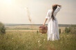 © junky_jess - Beautiful woman in vintage white dress with a big wicker basket with herbs standing in the middle of a meadow in summer, rear view, selective focus.