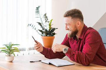 Successful relaxed red shirted freelancer discussing over video communication using phone while sitting at desk in office making notes in planner laughing merrily. Video conference with work partners