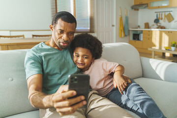 Wall Mural - Black African American father and son using smartphone at home. Happy smiling African Man and little child having video call