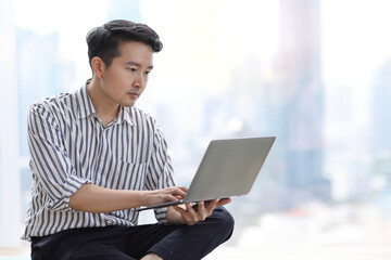 Wall Mural - Young asian business man sitting and using computer in business office building and city background