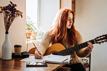 Unaltered Candid Portrait Of Young Red Haired Woman In Sweater Playing Acoustic Guitar Sitting By Window At Home. Hobbies, Indoor Home Activities For Adults In Winter, Autumn.