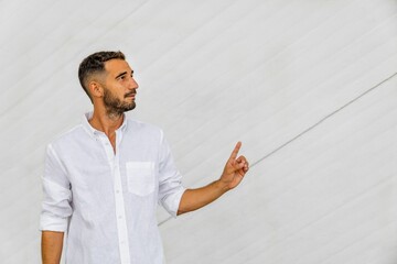 Hispanic man with beard facing away in white shirt pointing with hand on white concrete background.