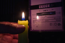 Burning Candle Near A Domestic Electric Meter In Darkness. Power Outage, Blackout, Load Shedding, Electricity Off Or Energy Crisis, Concept Image. 