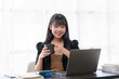 © amnaj - At work, a beautiful Asian business woman sit with her favorite cup of coffee enjoying a break at the office with her laptop.