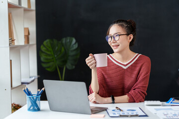 Wall Mural - At work, a beautiful Asian business woman sit with her favorite cup of coffee enjoying a break at the office with her laptop.