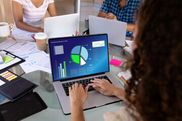 Wall Mural - Biracial businesswoman at desk in office using laptop with charts and data on screen