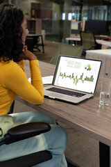 vertical of biracial businesswoman using laptop in empty office with graph and data on screen