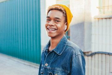 Young happy smiling latin american boy with curly afro hair listens music on headphones at street dressed in denim jacket and yellow beanie hat. Technology concept.