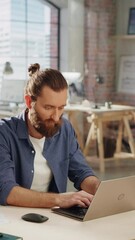 Poster - Vertical Screen: Handsome Long-Haired Bearded Manager Sitting at a Desk in Creative Office. Young Stylish Man Using Laptop Computer in Marketing Agency. Colleagues Working in the Background.