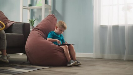 Adorable toddler boy plays video game on tablet computer sitting in bean chair at home. Development application for little children. Domestic lifestyle