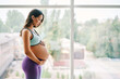 © GVS - Portrait of young pregnant woman in sportswear standing near window holding hands on her belly with copy space