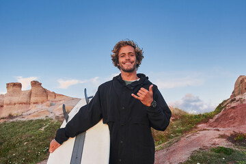 Wall Mural - Cheerful positive man surfer carrying his surfboard and showing shaka gesture to the camera