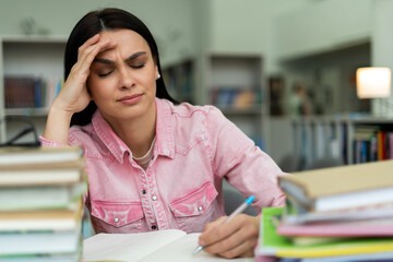 Waist up portrait view of the tired young woman feeling sleepy while reading a book and writing essay sitting at the table at the library