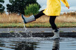 © okrasiuk - Woman having fun on the street after the rain. Cropped woman wearing rain rubber boots and yellow raincoat walking into puddle with water splash and drops. Fall weather. Selective focus