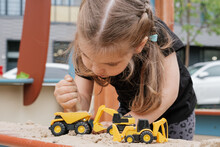 Little Girl Playing With Toy Trucks In Sandbox Outdoors