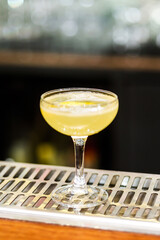 Glass of yellow cocktail decorated with lemon at bar counter background.