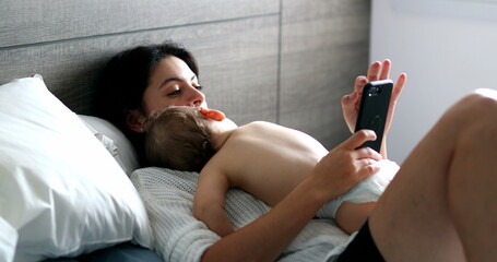 Wall Mural - Casual mother looking at cellphone with baby napping in bedroom, infant asleep on mom chest