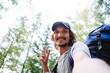 Cheerful selfie of a young guy against the background of the forest, a smile on his face, a gesture with his fingers, a hipster in a baseball cap, a hike in the mountains with a backpack