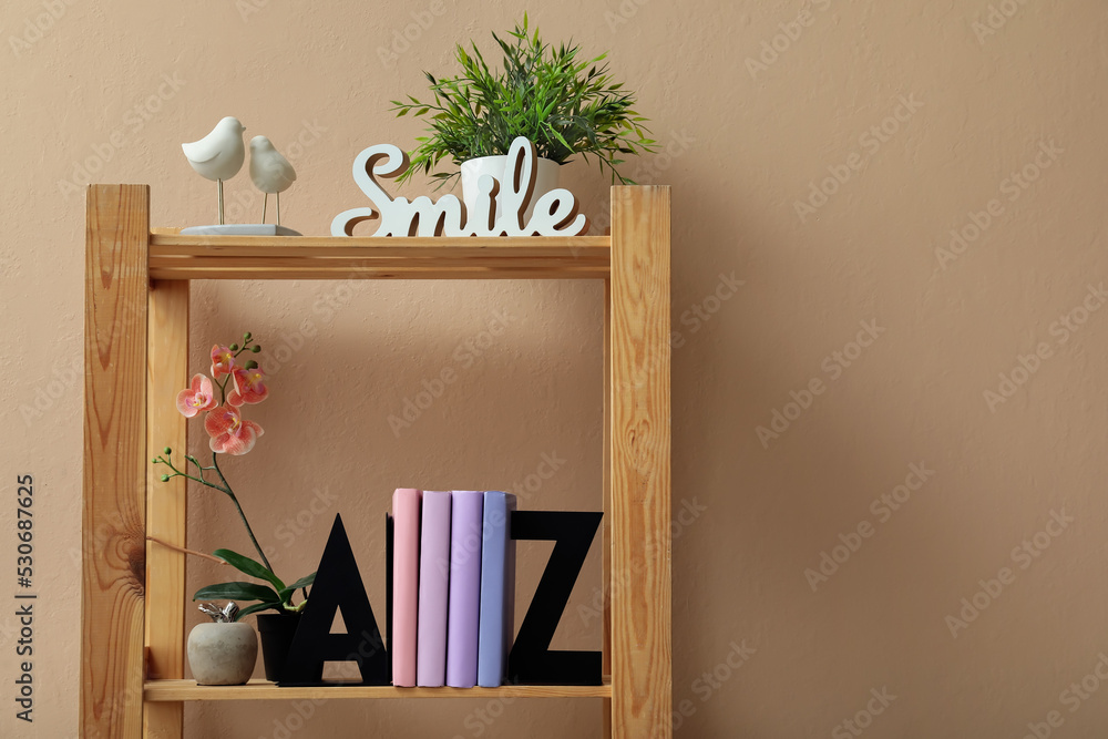 Bookcase with stylish holder, flowers and decor near beige wall