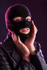 Wall Mural - Young woman in balaclava and leather jacket with hands near face against dark background