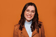 Closeup portrait of young carefree caucasian businesswoman smiling with positive expression, wears eyeglasses, blue turtle neck and brown jacket, stands over orange wall with blank space area.